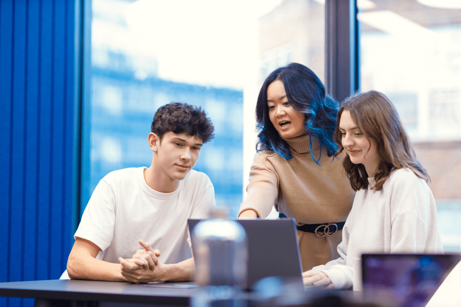 Um grupo de três pessoas reunidas em torno de um laptop. Uma mulher de moletom cor de bronze, com longos cabelos castanhos e mechas azuis, está explicando algo a dois colegas mais jovens. Um colega é homem e o outro é mulher; ambos estão usando blusas brancas.